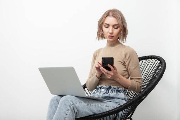Business serious woman millennial uses a smartphone and sits on an armchair with a laptop against the background of a white wall. Remote work - Photo, image