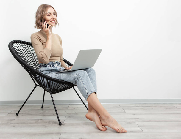 Üzleti mosolygós szőke nő telefonál és laptopot használ, miközben egy kényelmes karosszéken ül a nappaliban. Távmunka koncepció, életmód - Fotó, kép