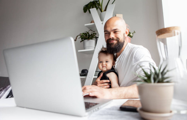 Feliz barbudo padre trabajador con hija pequeña utiliza el ordenador portátil mientras está sentado en la mesa en la habitación moderna - Foto, imagen
