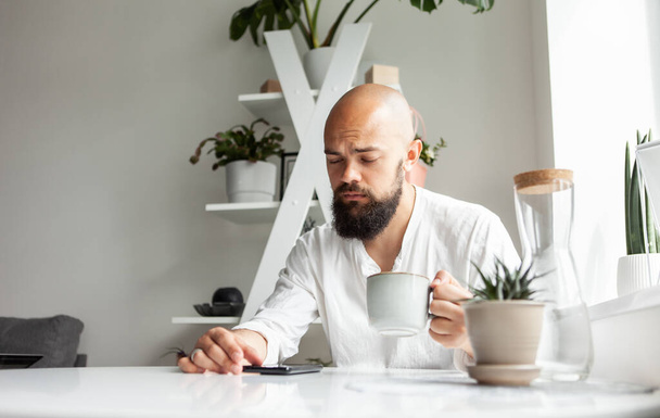 Handsome bald bearded man drinking coffee and using smartphone while sitting at the table in modern kitchen. Online work or leisure. Young male checks messages or reads news - Zdjęcie, obraz