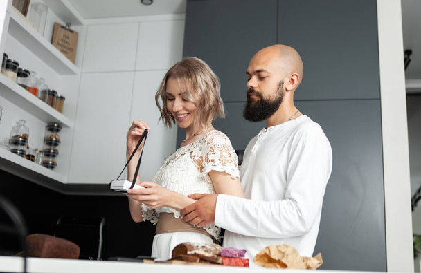 Handsome man makes a surprise to his lovely woman while she is cooking in the kitchen. The husband gives a box with a gift to his wife. Romantic love concept. Birthday or anniversary - Φωτογραφία, εικόνα