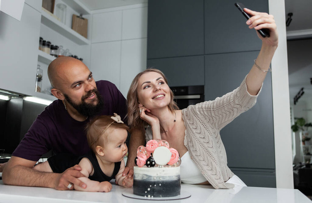 Anniversaire. Joyeux concept de famille. Jeune maman prend un selfie de sa fille et son mari avec un gâteau dans la cuisine - Photo, image