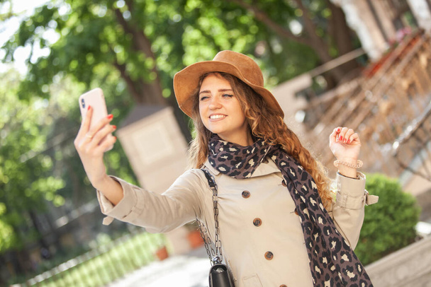 Cute smiling caucasian woman in hat and trench coat makes selfie in the city - Photo, image