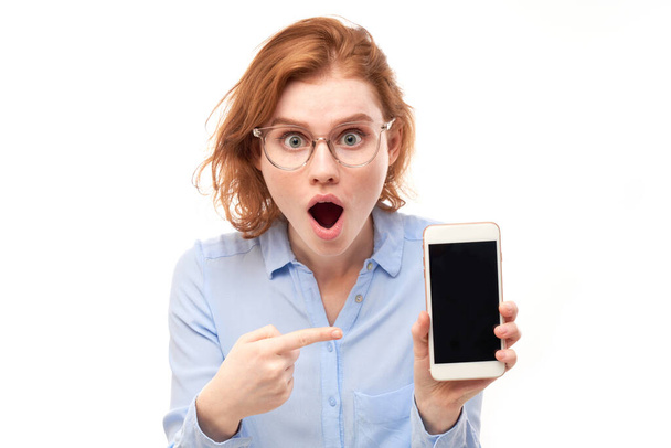 Shocked joyful redhead girl points finger at smartphone blank screen in glasses and business shirt isolated on white studio background - Фото, зображення