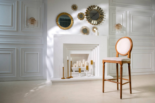старинный деревянный стул на фоне белого камина в стене с большими свечами и круглыми золотыми зеркалами - Фото, изображение