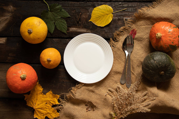 white plate stands on a wooden table next to pumpkins and autumn leaves, festive autumn table setting, dishes on the table - Photo, image