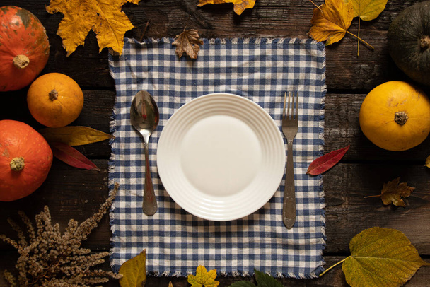 witte plaat op het tafelkleed staat op een houten tafel naast pompoenen en herfstbladeren, feestelijke herfsttafelopstelling, schotels op tafel - Foto, afbeelding