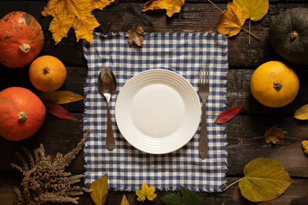 white plate on the tablecloth stands on a wooden table next to pumpkins and autumn leaves, festive autumn table setting, dishes on the table - Photo, Image