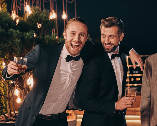 Two handsome men in suits drinking whiskey and smiling while spending time on party - Foto, Imagem