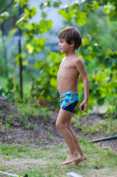 Παιδική εξωτερική δραστηριότητα. Χαμόγελο αγοράκι φορώντας ένα μπλε μαγιό σορτς τρέχει και παίζει με πιτσιλιές νερού διασκεδάζοντας σε μια αυλή σε μια ηλιόλουστη ζεστή μέρα του καλοκαιριού. Καυκάσιος παίζει κρυφτό - Φωτογραφία, εικόνα