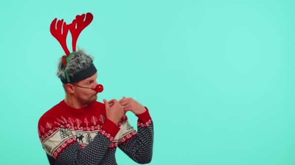 Mężczyzna nosi czerwony sweter noworoczny jeleń pokazując kciuki do góry i wskazując na pustą przestrzeń reklamową - Materiał filmowy, wideo