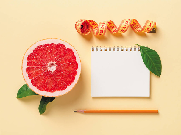 Half of ripe juicy grapefruit, roll of measuring tape, spring notepad and pensil on a pastel yellow background. Slimming diet, body weight control and healthy eating. Copy space. Top view. - Fotoğraf, Görsel