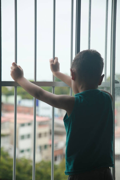 salvador, bahia, Brasile - 6 novembre 2021: bambino accanto a una griglia in una finestra di un appartamento durante un periodo di isolamento sociale a causa di coronavirus nella città di Salvador. - Foto, immagini