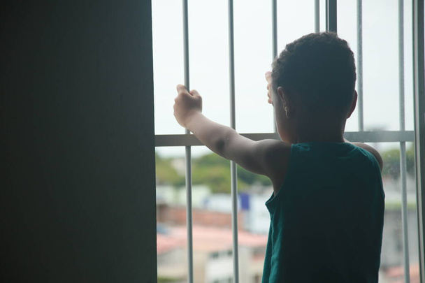 Сальвадор, Баия, Бразилия - 6 ноября 2021 года: ребенок рядом с решеткой в окне квартиры в период социальной изоляции из-за коронавируса в городе Сальвадор. - Фото, изображение