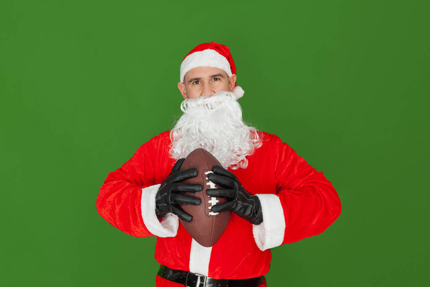 A Caucasian man dressed as Santa Claus looking straight ahead holding a brown american football ball in his hands. The background is green. - Photo, image