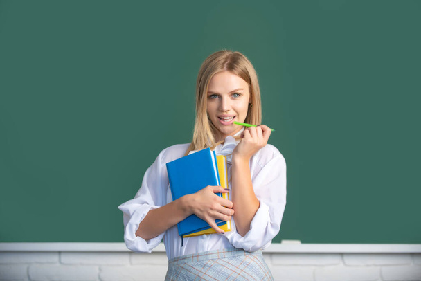 Portrait of young female college student studying in classroom on class with blackboard background. Creative young smiling female student with pen and book. - Foto, afbeelding