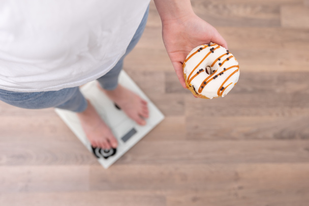 A woman stands on the scales, holds a donut in her hands, top view, copy space. - Fotografie, Obrázek