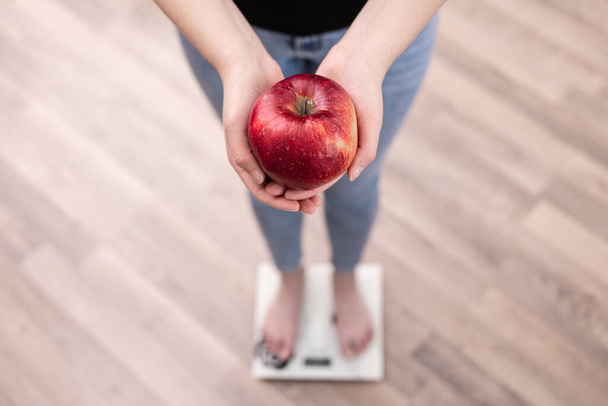 Μια γυναίκα μετράει το βάρος σε μια κλίμακα, κρατά ένα μήλο στα χέρια της. - Φωτογραφία, εικόνα