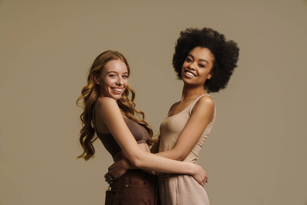 Two smiling diverse young women in underwear standing over beige background hugging - Foto, Imagem