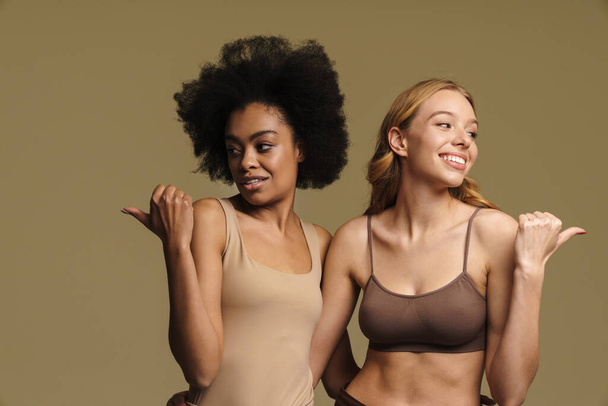Two diverse smiling young women in underwear standing over beige background pointing finger at copy space - Fotografie, Obrázek