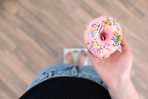 Een vrouw staat op de weegschaal, houdt een donut in haar handen, bovenaanzicht, kopieerruimte. - Foto, afbeelding