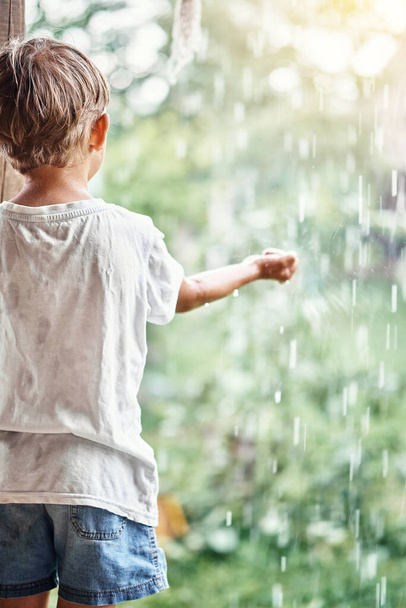 Blond mały chłopiec w białym t-shirt trzyma rękę pod spadające krople deszczu stojąc na tarasie wiejskiego domu widok z tyłu - Zdjęcie, obraz