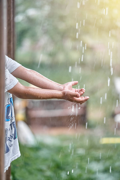Blond little boy in white t-shirt holds hand under falling down rain drops standing on country house terrace backside view - Foto, Imagen