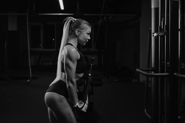 Een foto van de zijkant van een fitte vrouw met lang blond haar die een triceps touw duwt in een sportschool. Een meisje traint haar armen.. - Foto, afbeelding