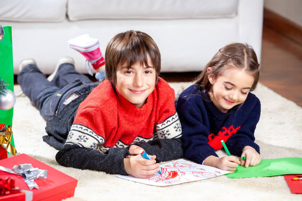 Zwei kleine Kinder liegen auf dem Fußboden ihres Hauses und schreiben Weihnachtsbotschaften - Foto, Bild