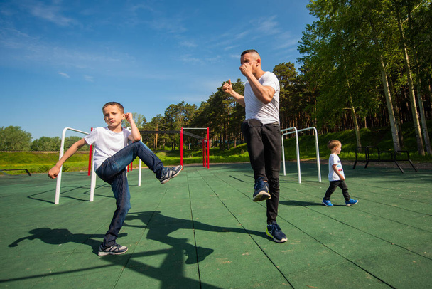 Hombre caucásico y dos chicos haciendo ejercicios al aire libre. Padre e hijos entrenan en el campo de deportes - Foto, imagen