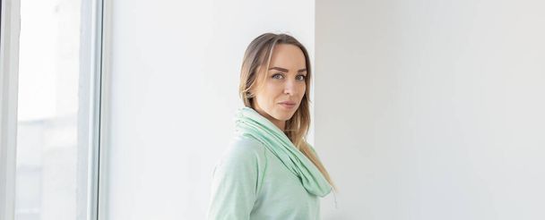 Portrait of cute positive woman in mint hoodie standing near window relaxing in living room at her home. Copy space and space for advertising - Photo, Image