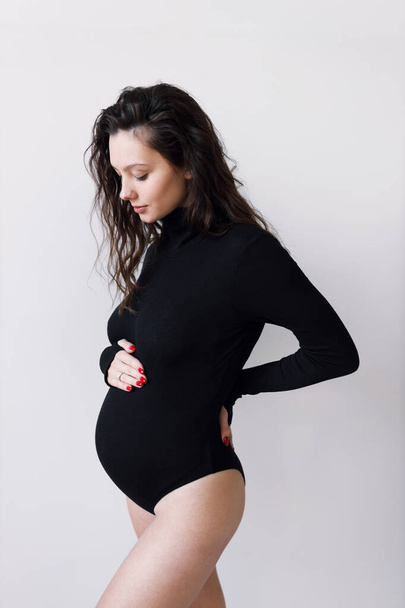 The concept of a fashionable image for pregnant women. A young beautiful pregnant woman in a black bodysuit, on a white wall. - Foto, Bild