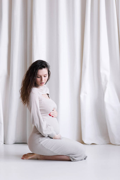 A concept for a pregnant stylish girl. A young beautiful pregnant woman in a white dress, on a light background. - 写真・画像