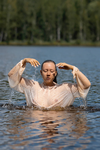 Tender seductive female in white wet transparent shirt standing in water of pond and looking away - Photo, Image