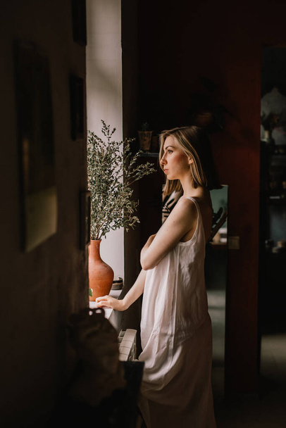 talented sculptor girl in light negligee stands in pottery workshop near large window. concept is beauty - Photo, Image