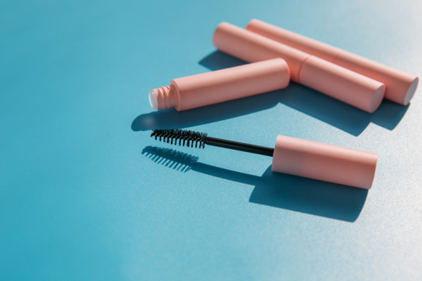 escova de maquiagem rosa limpa para rímel fica ao lado de um tubo aberto, tubos fechados de cosméticos, brilho labial, batom líquido, delineador rosa em um fundo azul com sombras. Espaço de cópia - Foto, Imagem