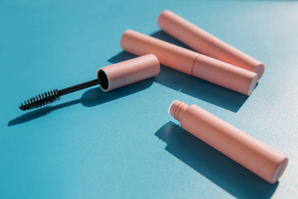 pink clean makeup brush for mascara lies next to an open tube, closed tubes of cosmetics, lip gloss, liquid lipstick, pink eyeliner on a blue background with shadows. Copy space - Photo, Image