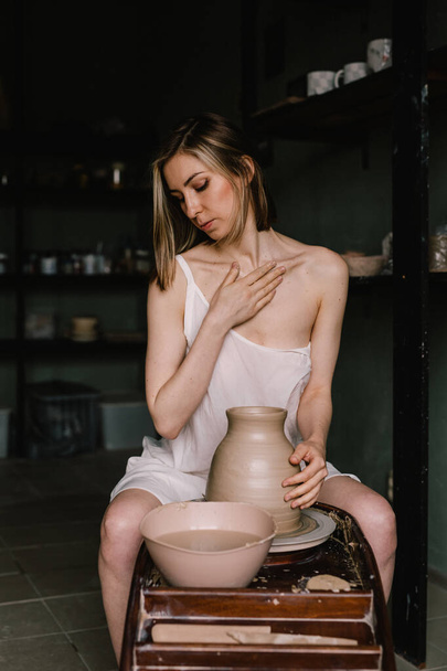 talented sculptor girl in thin light negligee sculpts a jug of clay in pottery workshop near large window. Concept of sculpture - Zdjęcie, obraz