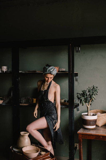 beautiful sculptor girl with bandage on her head and a black apron on her naked body is standing in pottery workshop near large window. concept is beauty - Photo, Image