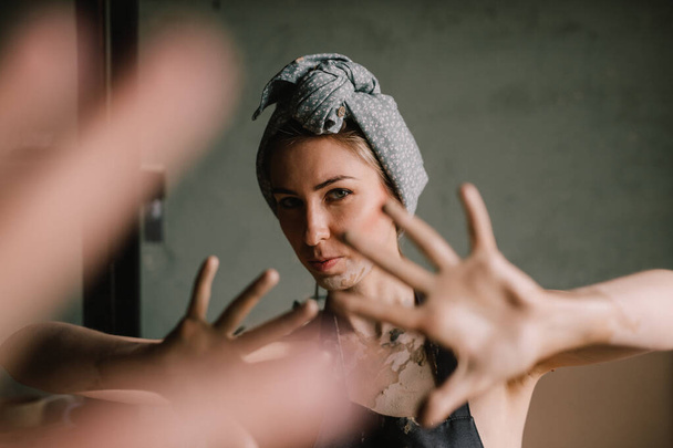 beautiful sculptor girl with a headband and a black apron shows her hands stained with clay. camera focus is on girl's face, blur is on her hands. concept is beauty and art. - Photo, Image