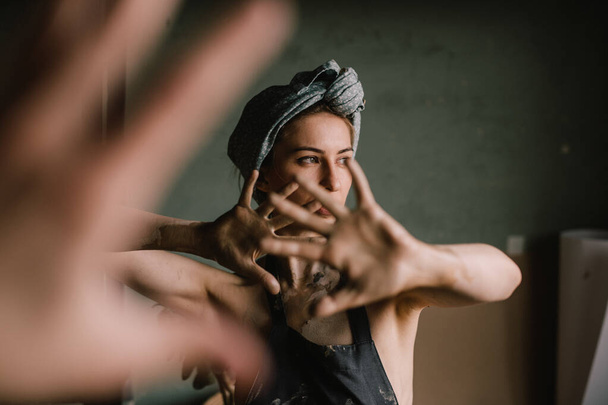 beautiful sculptor girl with a headband and a black apron shows her hands stained with clay. camera focus is on girl's face, blur is on her hands. concept is beauty and art. - Фото, изображение