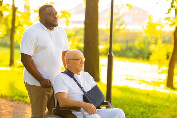 Caregiver and old man in a wheelchair. Professional nurse and patient walking outdoor in the park at sunset. Assistance, rehabilitation and health care. - Φωτογραφία, εικόνα