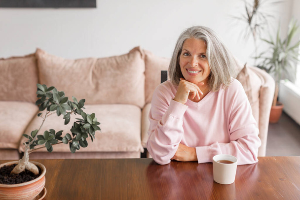 Woman have relax at home with cup of tea. Happy gray-haired middle aged female at home - 写真・画像