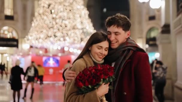 Jonge volwassen man met bloemen kussen en knuffelen zijn vrouw op de achtergrond van de kerstboom. Gelukkig paar in de liefde knuffels nadat ze ja zei in de Vittorio Emanuele II galerie winkelcentrum in Milaan, Italië - Video