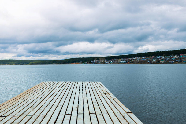 an empty pier in the foreground, a pond with a pier and a village. cloudy sky in the distance forest - Photo, Image