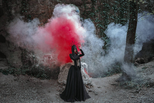 Νεαρή γυναίκα με μάγισσα στέκεται με μαύρο φόρεμα και στέμμα στο κεφάλι της σε σκοτεινό και ομιχλώδες δάσος. Αποκριάτικη στολή για σκοτεινό πάρτι. - Φωτογραφία, εικόνα