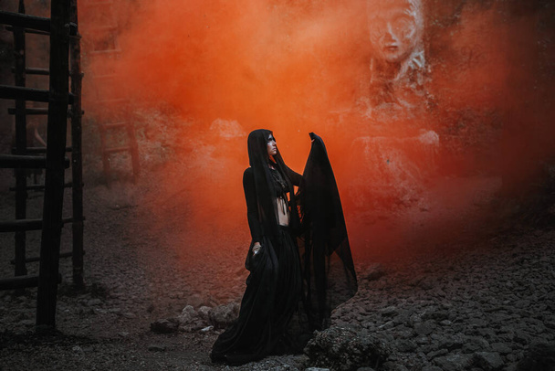 Jonge vrouw in heks staat in zwarte jurk in donker en mistig bos. halloween kostuum voor donker feest. - Foto, afbeelding