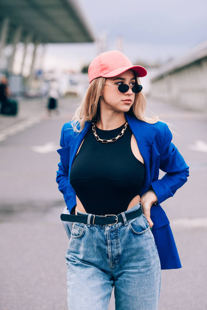 Jonge sexy blonde hipster vrouw poseren op straat. Het dragen van blauwe stijlvolle jas, jeans en honkbalpet en zonnebril. - Foto, afbeelding