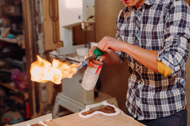 Timmerman steekt een brander aan in een werkplaats. Proces van het maken van een ambachtelijke hars en houten tafel - Foto, afbeelding