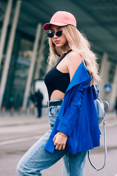 Young sexy blonde hipster woman posing on the street. Wearing blue stylish jacket, jeans and baseball hat and sunglasses. - Фото, изображение